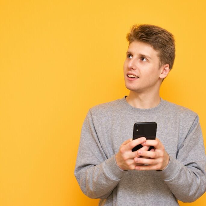 Portrait of a positive young man in the casual clothing stands with a smartphone in his hands, smiles and looks sideways on a yellow background. Copyspace